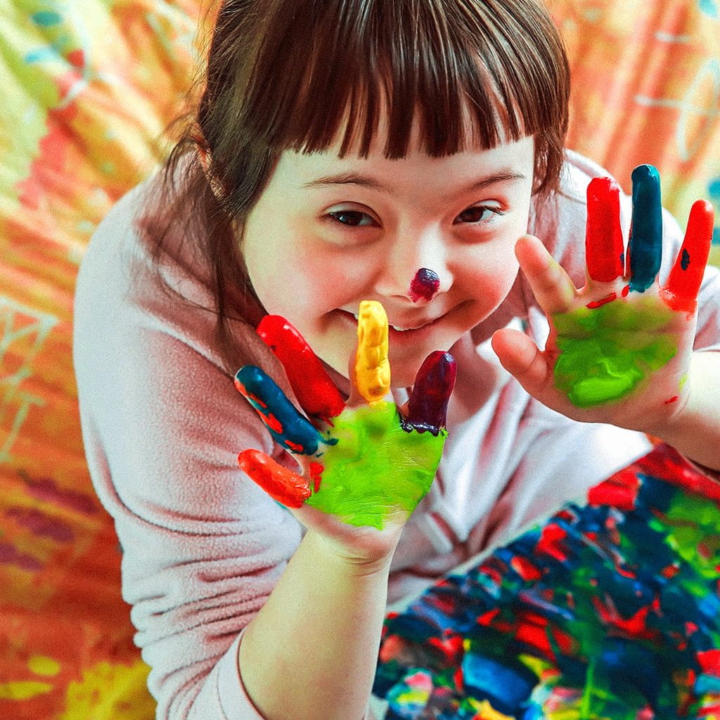 Ein Mädchen mit Trisomie 21 strahlt in die Kamera während sie mit Fingerfarben spielt