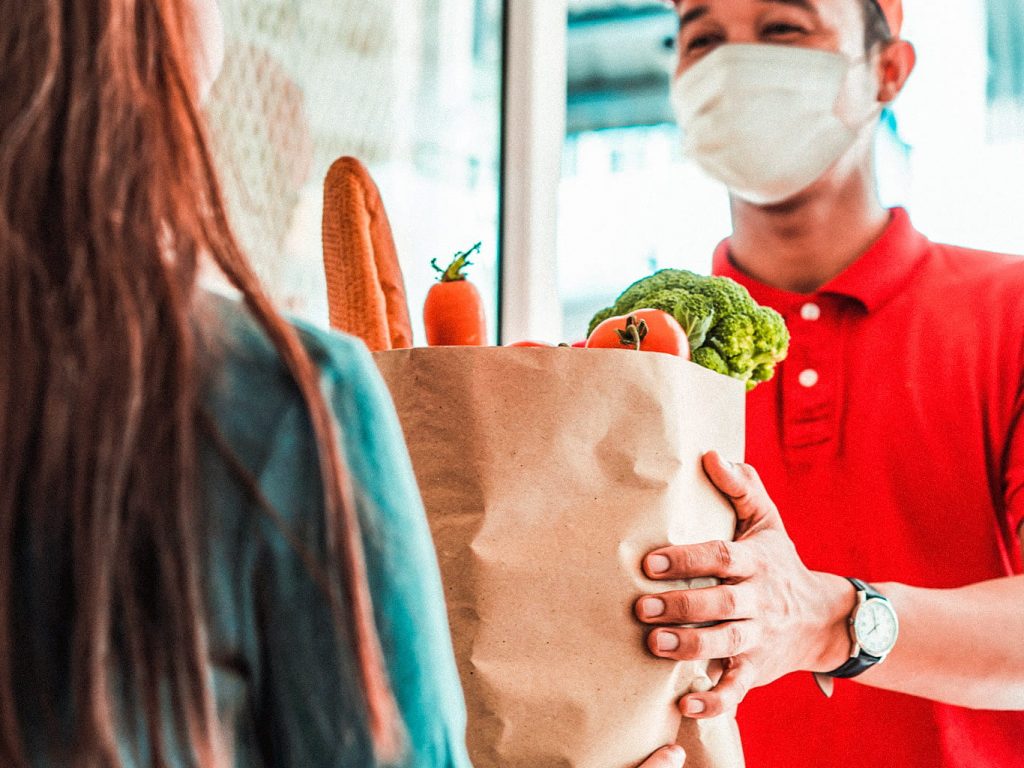 Ein Lieferant bringt einer Frau Lebensmittel zur Tür. Er trägt eine OP Maske