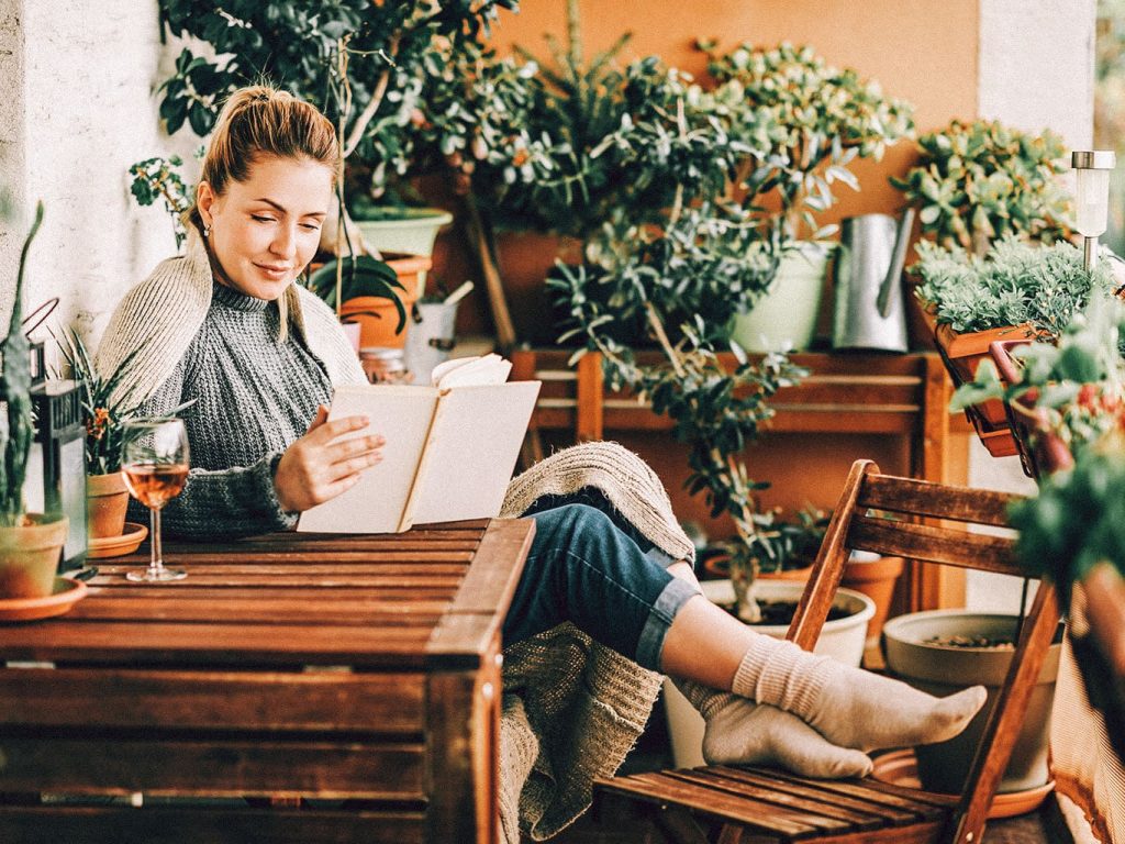 Eine Frau mit einem warmen Pullover an sitzt auf einem Gartenstuhl, liest ein Buch und trinkt dabei ein Glas Wein