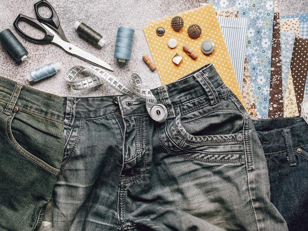 Ein paar alte Jeans sowie Nähwerkzeug und eine Schere liegen mit einigen Stoffrestens auf einer grauen Oberfläche