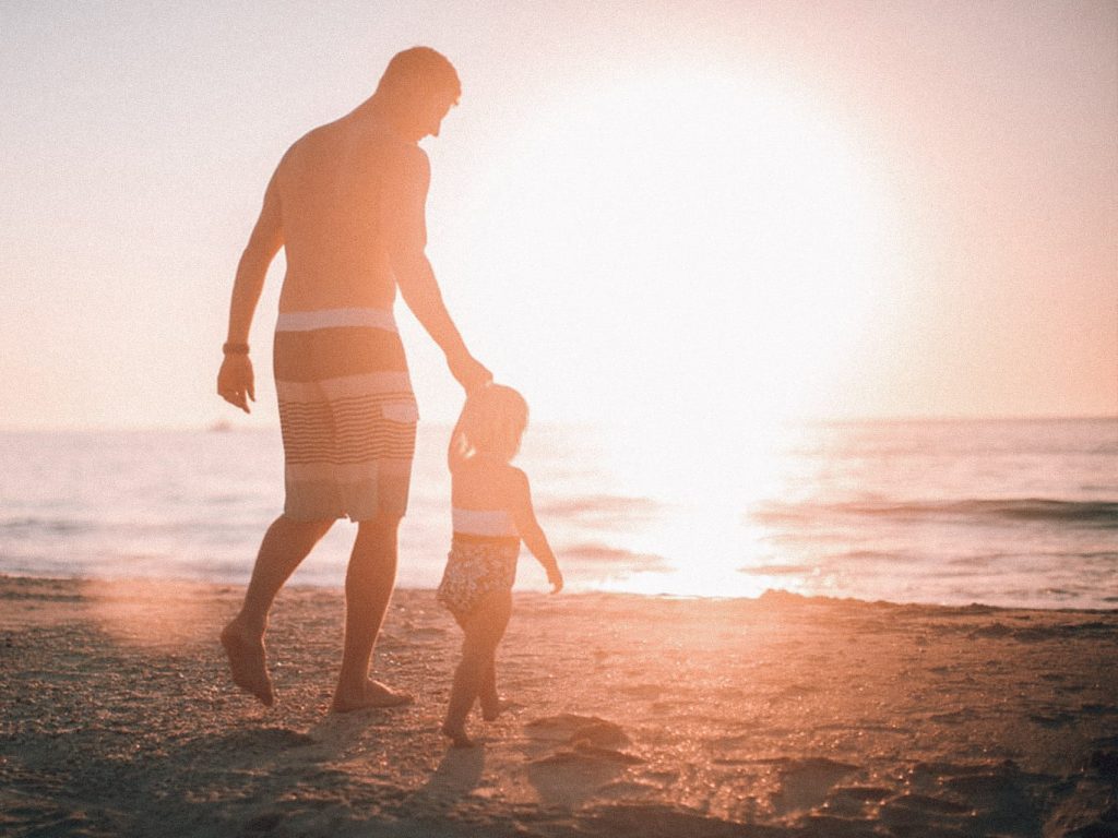 Ein Mann hält ein Kind an einer Hand. Gemeinsam gehen sie auf Sand in Richtung Meer