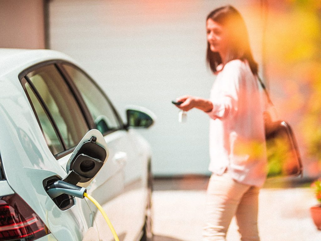 Eine Frau schließt ihr weißes Elektroauto ab, das an einer Ladesäule hängt