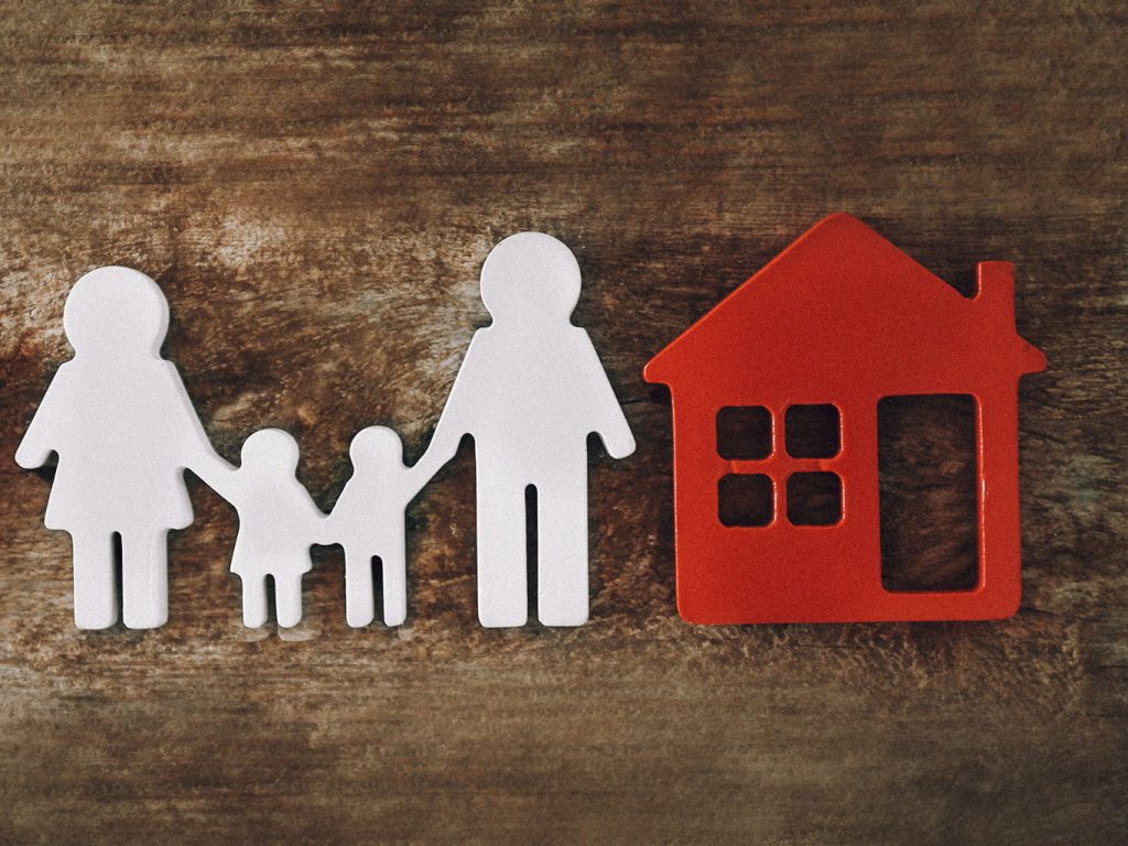 Eine Familie aus Vater, Mutter und zwei Kindern als Holzspielfiguren stehen neben einem roten Haus