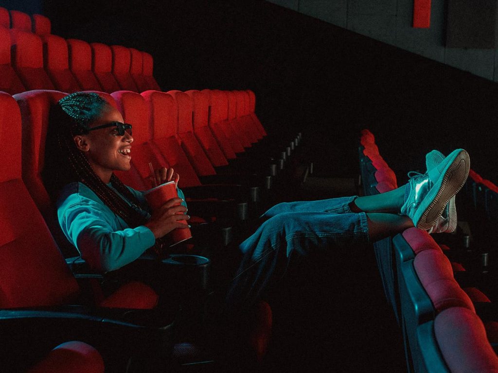Eine Frau sitzt mit ausgestreckten Füßen auf einem Kinosessel mit Popcorn in der Hand und lacht