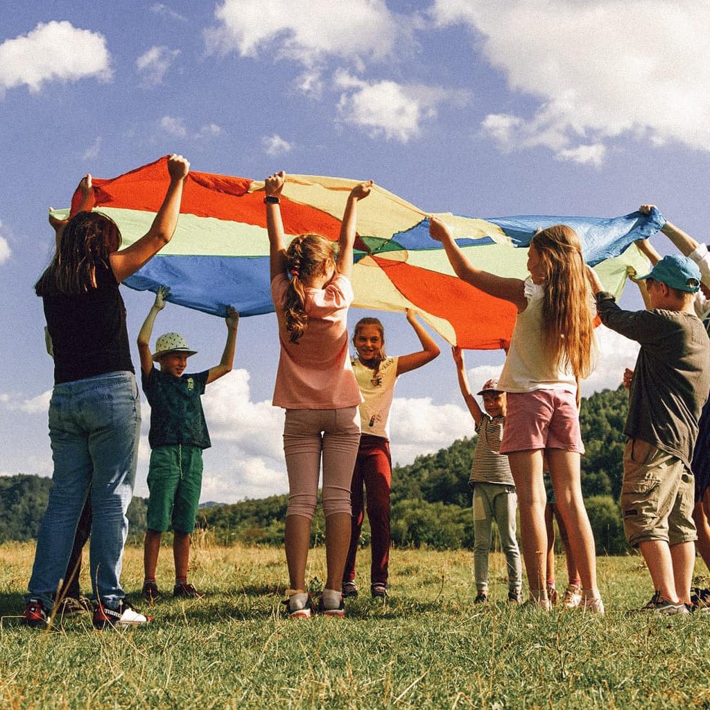 Eine Gruppe von Kindern steht im Kreis und alle halten ein großes Tuch in Regenbogenfarben hoch