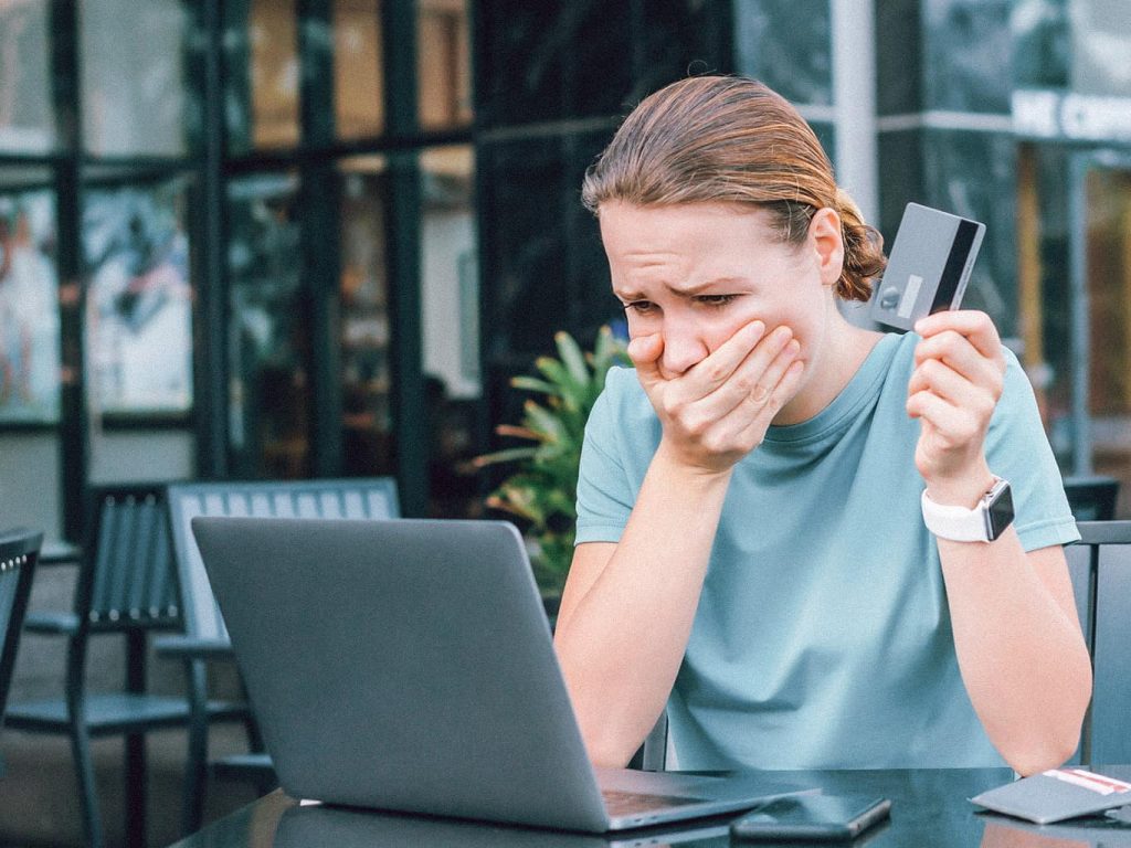 Eine Junge Frau sitzt mit der Kreditkarte in der Hand geschockt vor ihrem Laptop und hält sich mit der anderen Hand den Mund zu