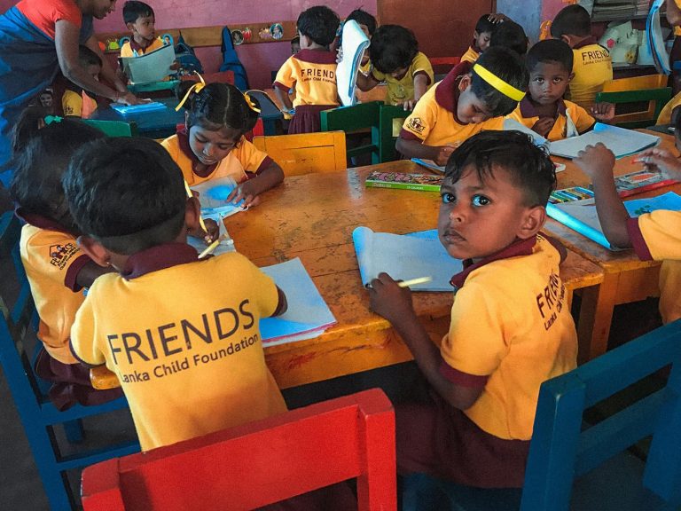 Mehrere Kinder mit T-Shirts der FRIENDS Kinderhilfe International Sri Lanka sitzen in einem Raum an einem Tisch und lernen das Schreiben