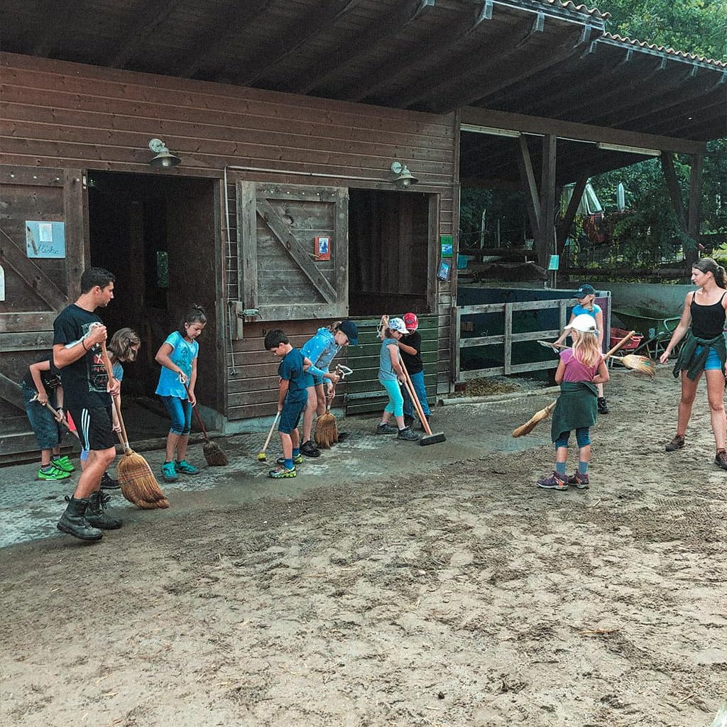 Eine Gruppe von kleinen Kindern und zwei Erwachsenen putzen mit Besen und Rechen den Boden vor einem Reitstall