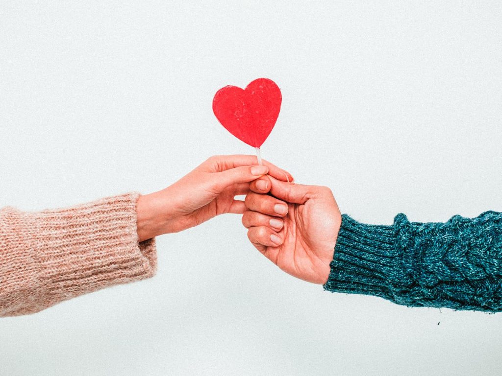Zwei Hände strecken sich zur Mitte und halten dort gemeinsam ein rotes Herz in den Händen.