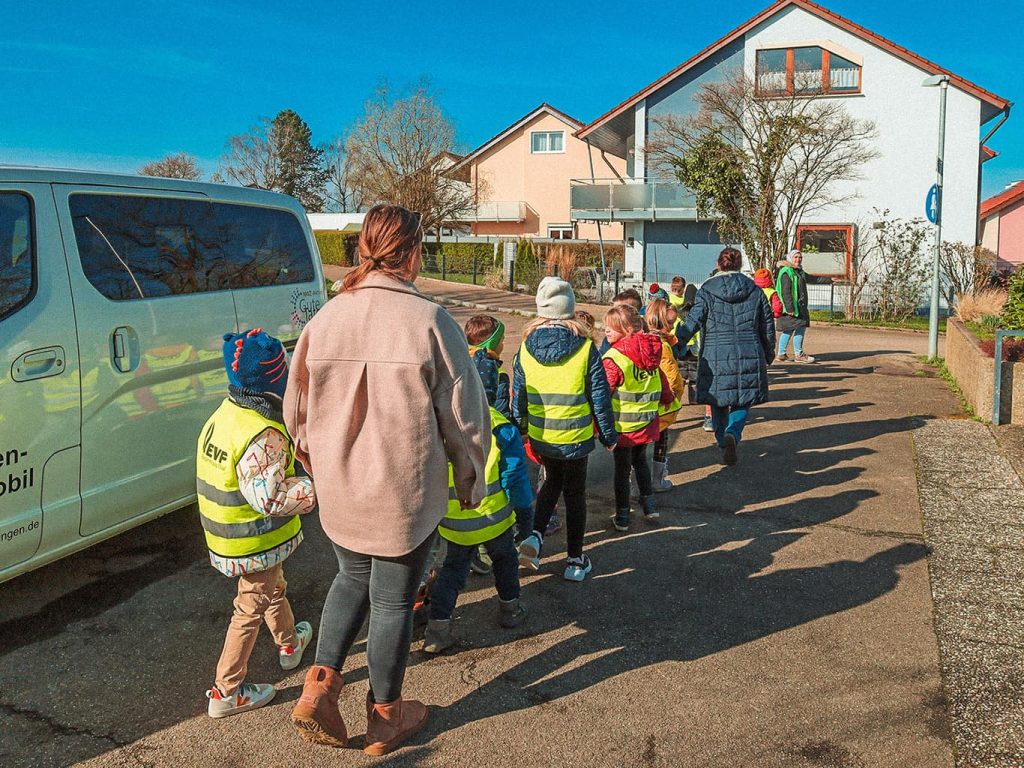 Eine Gruppe von Kindern in gelben Warnwesten werden von zwei Erwachsenen begleitet und gehen über eine Straße