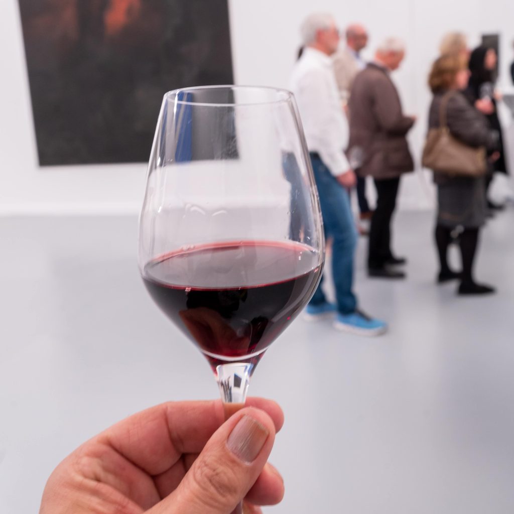 Eine Hand hält ein Glas Rotwein in die Kamera. Im Hintergrund ist eine Menschengruppe zu sehen.