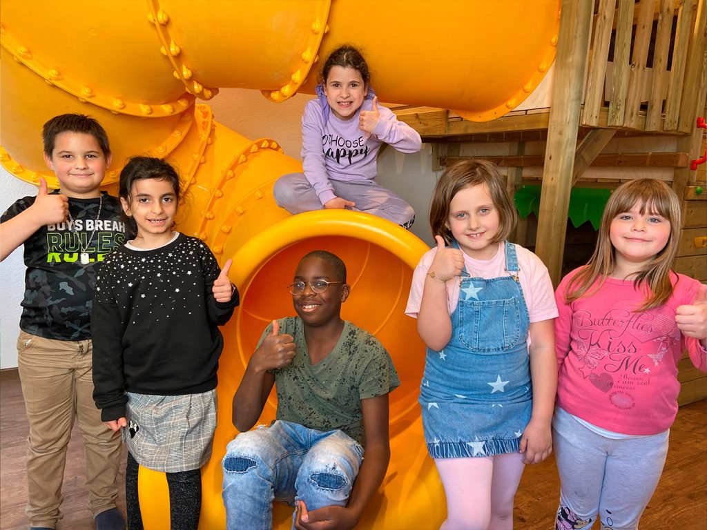 Eine Gruppe von Kindern steht und sitzt um eine gelbe Rutsche und halten ihre Daumen hoch