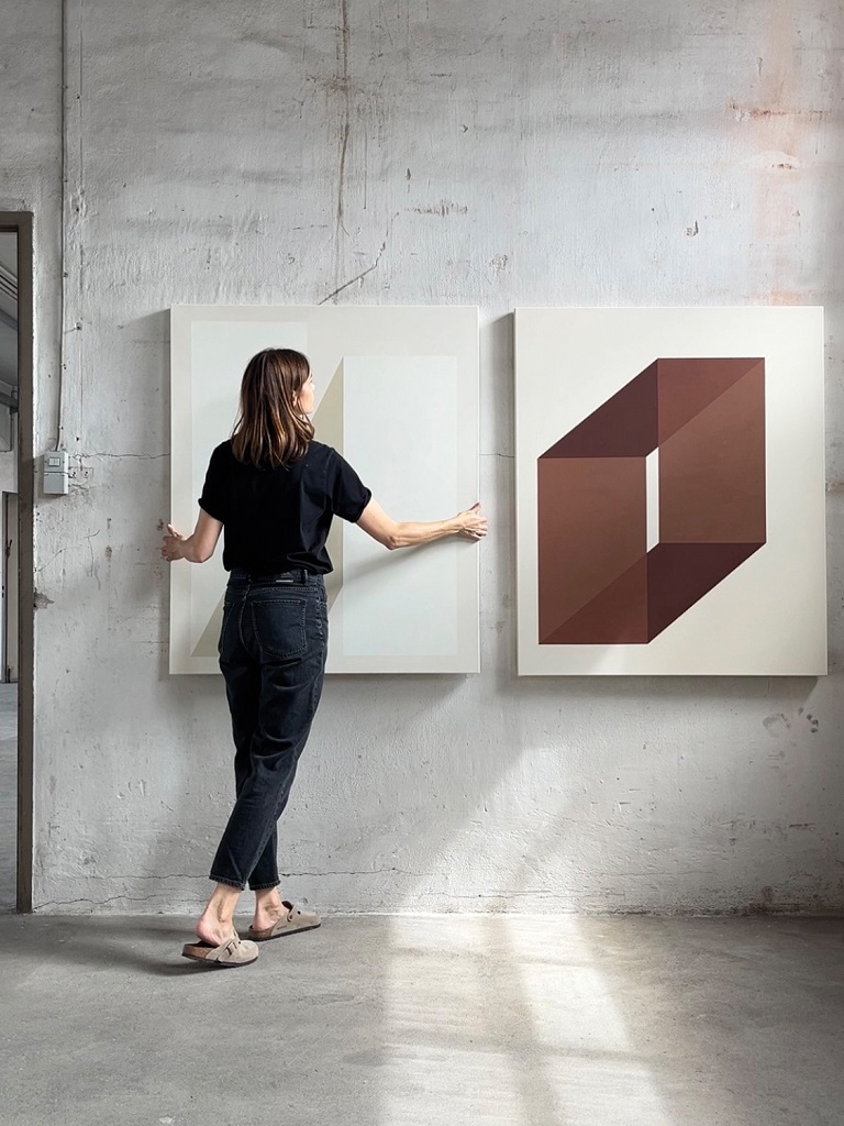 Carolin Kreutzer in einer lichtdurchfluteten Galerie, während sie zwei ihrer abstrakten, geometrischen Kunstwerke anordnet. Die Werke zeigen klare Linien und reduzierte Farbflächen in Weiß- und Brauntönen.