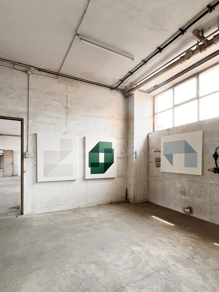 Ausstellungsraum mit drei großformatigen, abstrakten Kunstwerken von Carolin Kreutzer. Geometrische Formen in dezenten Grau- und Grüntönen hängen an einer rohen, industriellen Wand und erzeugen eine minimalistische, moderne Atmosphäre.