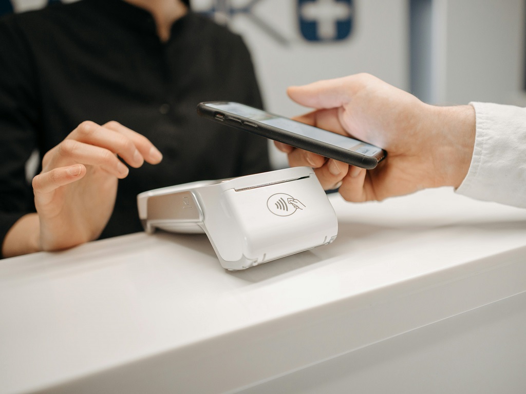 Eine Person nutzt digitales Banking und bezahlt kontaktlos mit einem Smartphone an einem weißen Kartenlesegerät, während eine Kassiererin im Hintergrund wartet.