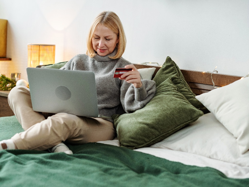 Eine Frau sitzt entspannt auf einem Bett, nutzt ein Laptop für digitales Banking und hält eine Bankkarte in der Hand.