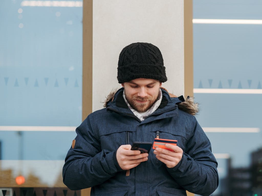 Ein Mann mit schwarzer Strickmütze und dunkelblauer Winterjacke steht draußen vor einer Glasfassade. Er hält in einer Hand ein Smartphone und in der anderen eine Bankkarte, während er sich auf das digitale Banking konzentriert.