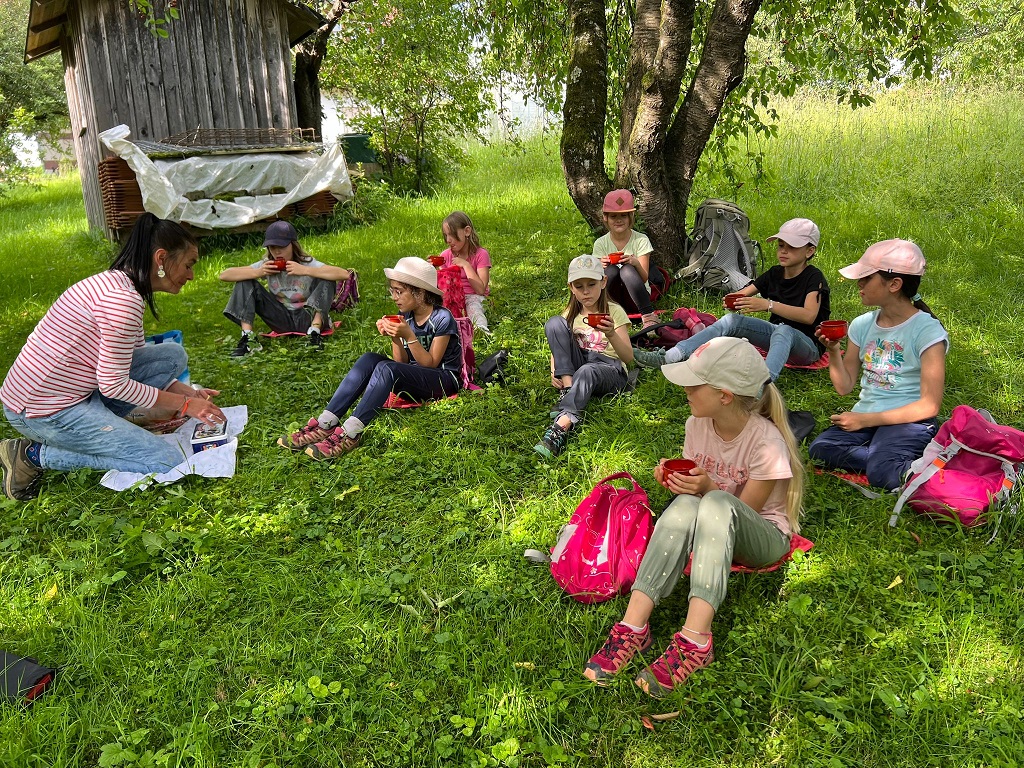 Eine Gruppe von Kindern sitzt auf einer Wiese unter einem schattigen Baum und hält rote Schalen in den Händen. Eine erwachsene Frau mit gestreiftem Shirt und Jeans kniet vor ihnen und zeigt ihnen etwas auf einem Blatt Papier. Die Kinder tragen sportliche Kleidung und Kappen, ihre Rucksäcke liegen neben ihnen im Gras. Im Hintergrund steht eine kleine Holzhütte. Die Szene wirkt naturverbunden und entspannt.