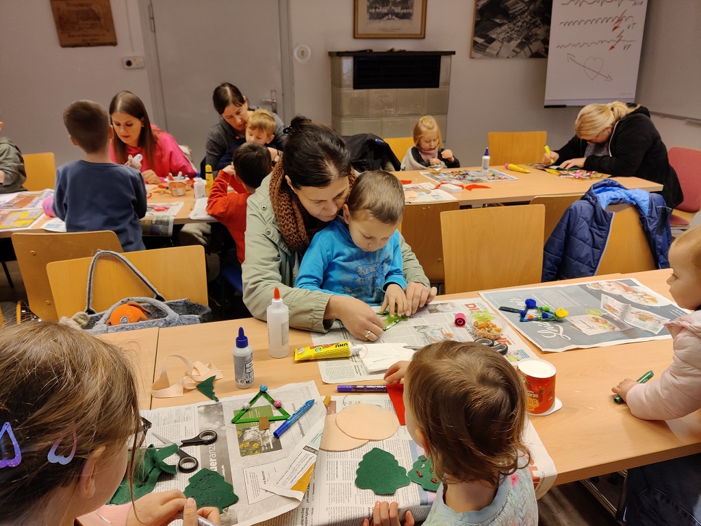 Mehrere Kinder und Erwachsene sitzen an Tischen in einem Raum und basteln gemeinsam mit verschiedenen Materialien wie Filz, Schere und Kleber. Eine Frau hilft einem Jungen konzentriert bei seiner Bastelarbeit. Auf den Tischen liegen Zeitungen als Unterlage. Im Hintergrund arbeiten weitere Kinder und Erwachsene an ihren Projekten. Die Atmosphäre wirkt warm und gemeinschaftlich.