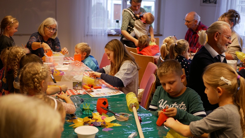 Kinder und Großeltern sitzen gemeinsam an Tischen und basteln bunte Laternen aus Transparentpapier und anderen Materialien. In der lebhaften und kreativen Atmosphäre helfen ältere Teilnehmende den Jüngeren. Der Raum ist mit Bastelmaterialien gefüllt, während im Hintergrund weitere Erwachsene und Kinder engagiert mitarbeiten.