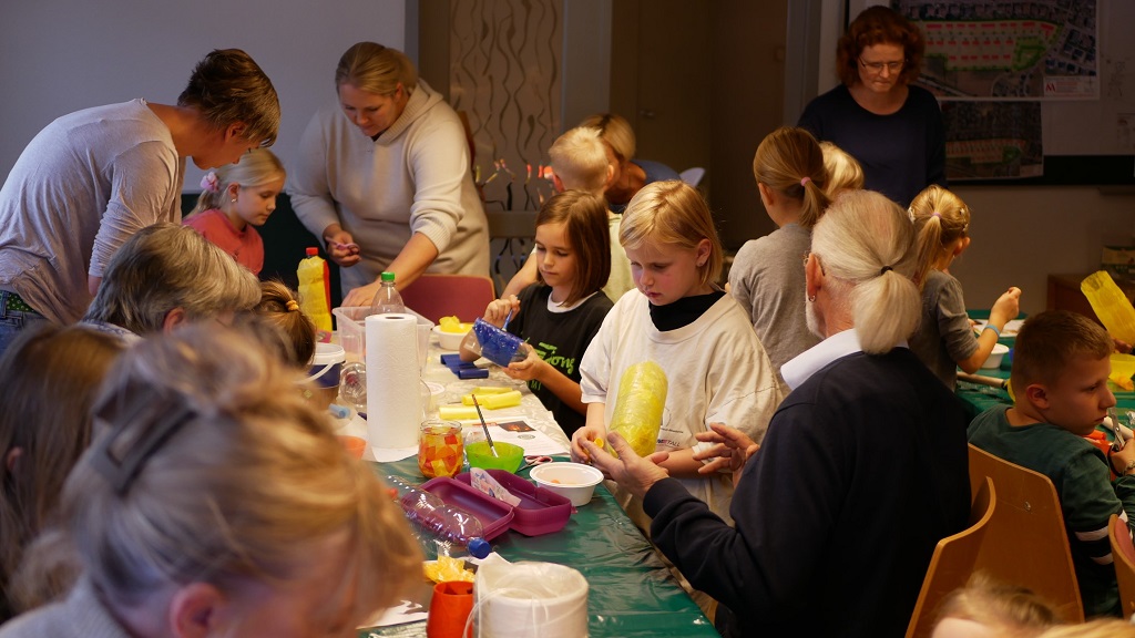Kinder und Erwachsene sitzen gemeinsam an Tischen und basteln bunte Laternen aus Transparentpapier und anderen Materialien. In der lebhaften und kreativen Atmosphäre helfen ältere Teilnehmende den Jüngeren. Der Raum ist mit Bastelmaterialien gefüllt, während im Hintergrund weitere Erwachsene und Kinder engagiert mitarbeiten.