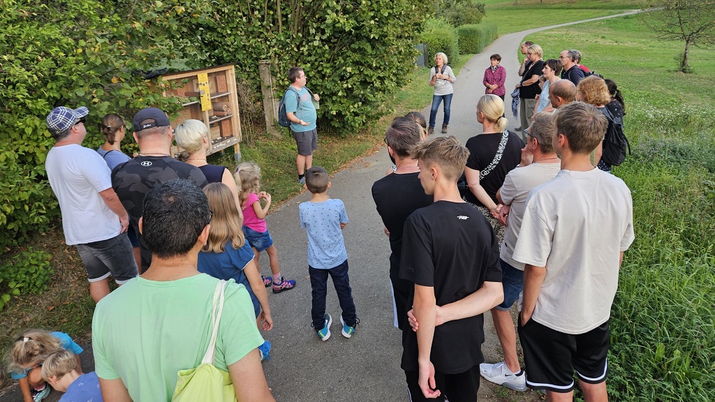 Eine Gruppe aus Erwachsenen und Kindern steht auf einem Weg in der Natur und hört einem Mann im blauen Shirt zu, der vor einem Insektenhotel etwas erklärt. Die Umgebung ist von grünen Wiesen und Büschen geprägt.