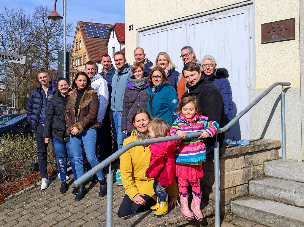 Eine Gruppe von Männern, Frauen und Kindern steht auf der Treppe vor einem historischen Gebäude und lächelt in die Kamera. Sie tragen warme Kleidung, es ist ein sonniger Tag. Im Hintergrund sind Häuser mit Solarpanelen auf den Dächern zu sehen.