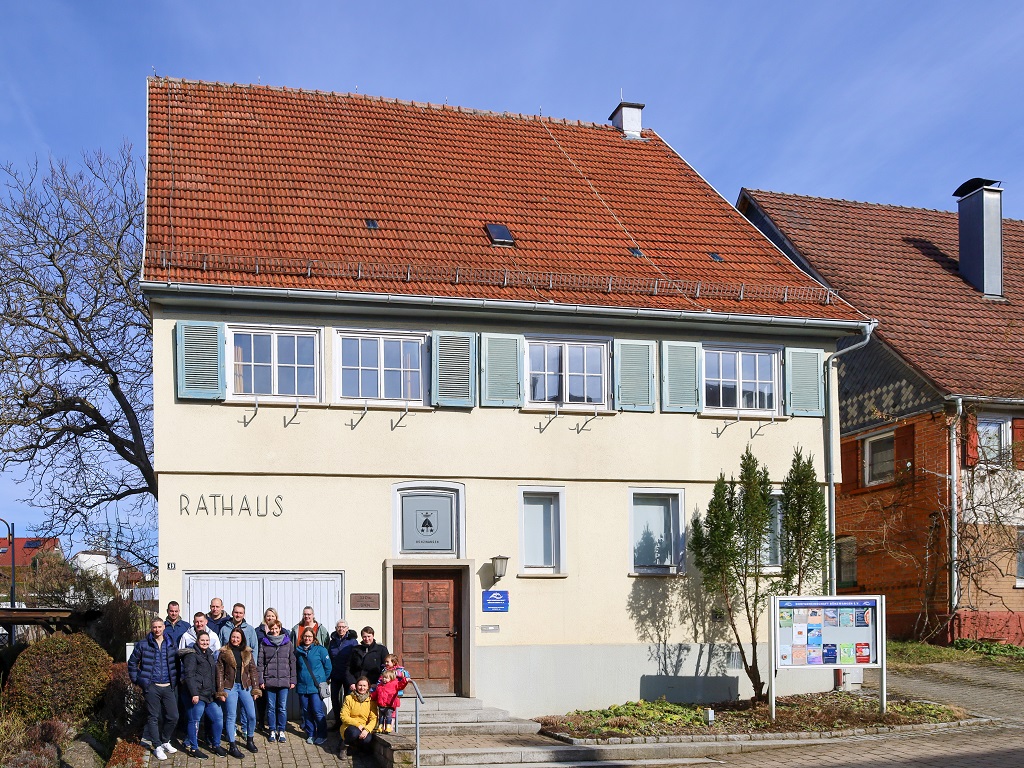 Das historische Rathaus Bünzwangen, ein zweigeschossiges Gebäude mit hellen Wänden, blauen Fensterläden und einem roten Ziegeldach. Vor dem Eingang steht eine Gruppe engagierter Bürger, die sich für den Erhalt des Gebäudes einsetzt. Rechts neben dem Rathaus befindet sich eine Infotafel.