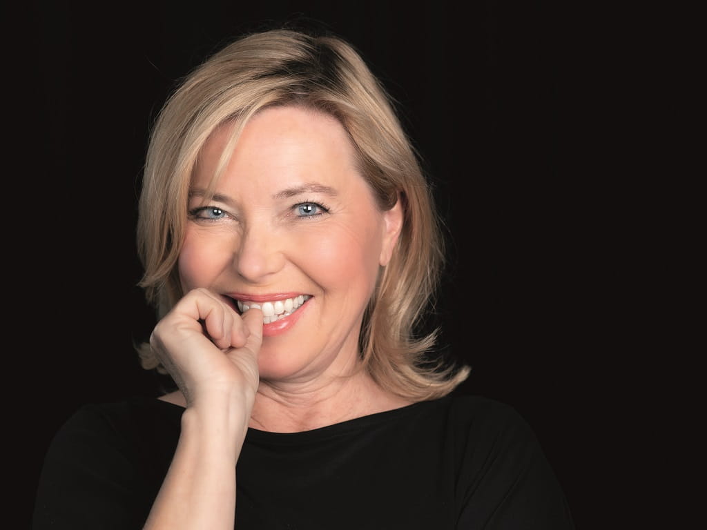 Porträt der Kabarettistin Simone Solga beim Süßener Kulturfrühling 2026 vor dunklem Hintergrund, lächelnd mit leicht schräg geneigtem Kopf und Hand am Kinn.