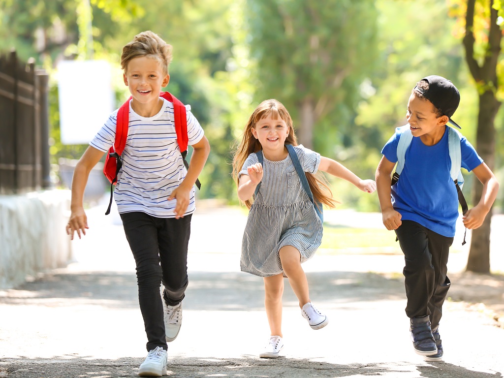 Drei Grundschulkinder mit Rucksäcken laufen lachend auf einem sonnigen Schulweg – ein fröhlicher Moment im Rahmen des SpoSpiTo Bewegungs-Passes für mehr Bewegung und einen autofreien Schulweg.