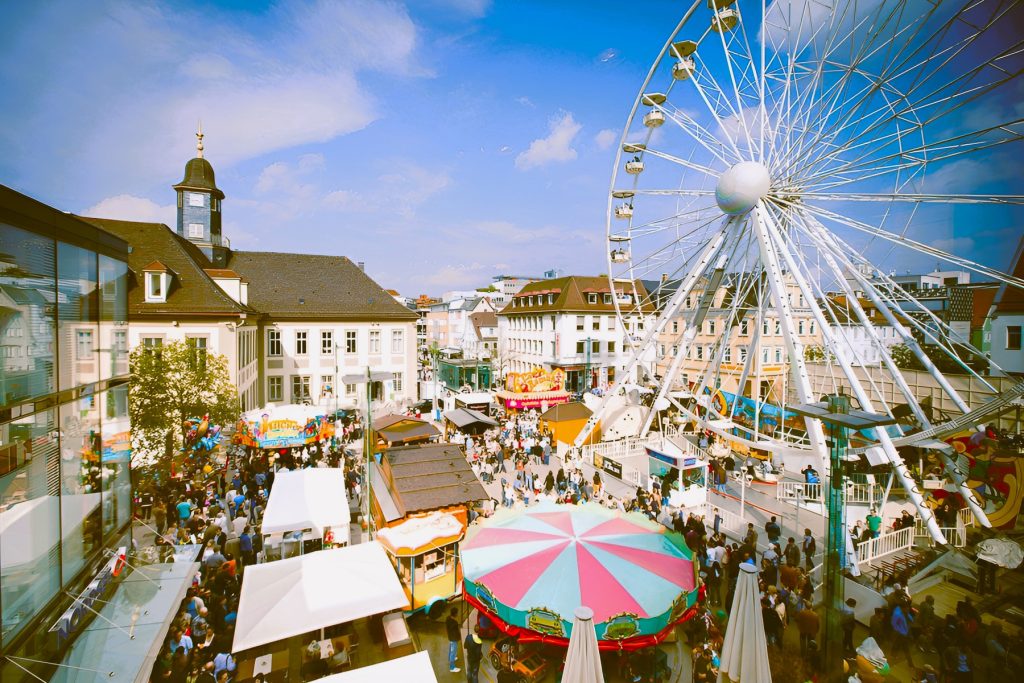 Das Frühlingsfest „Göppinger Frühling 2025“ in der Innenstadt mit dem großen Riesenrad „White Star“ als Blickfang. Besucher schlendern über den Festplatz mit bunten Marktständen, Fahrgeschäften und einem nostalgischen Karussell. Historische und moderne Gebäude rahmen die lebhafte Szenerie ein.