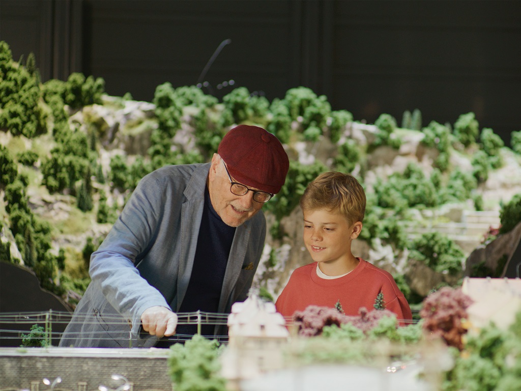 Älterer Mann mit roter Mütze zeigt einem Jungen eine detailreiche Modelleisenbahn-Anlage im Märklineum, umgeben von Miniatur-Landschaften.