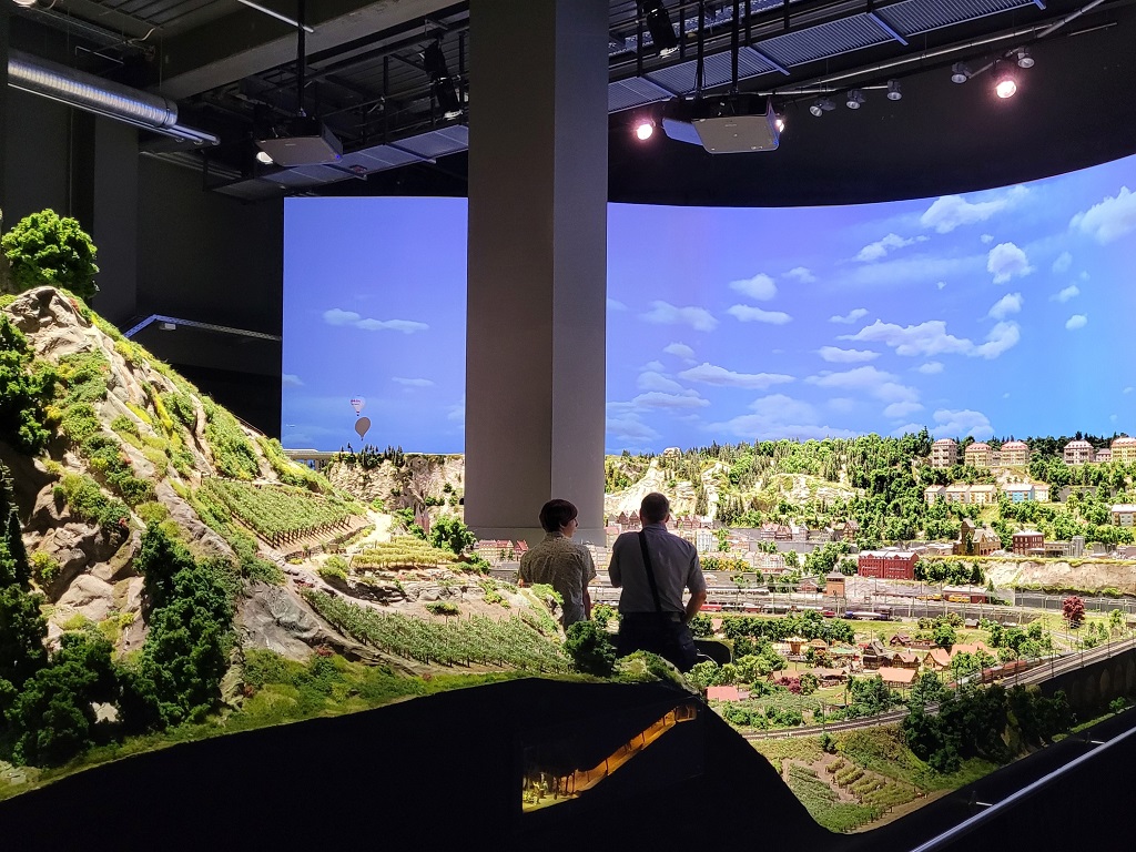 Zwei Besucher stehen vor einer großen Modelleisenbahnlandschaft im Märklineum Göppingen mit detailreicher Szenerie und digitalem Tag-Nacht-Himmel.