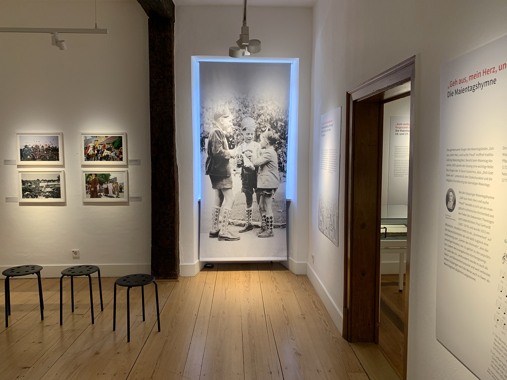 Ausstellungsraum mit historischen und farbigen Maientagsfotos, großformatigem Kinderfoto, Texttafeln zur Maientagshymne und mehreren Sitzhockern im Stadtmuseum Göppingen.
