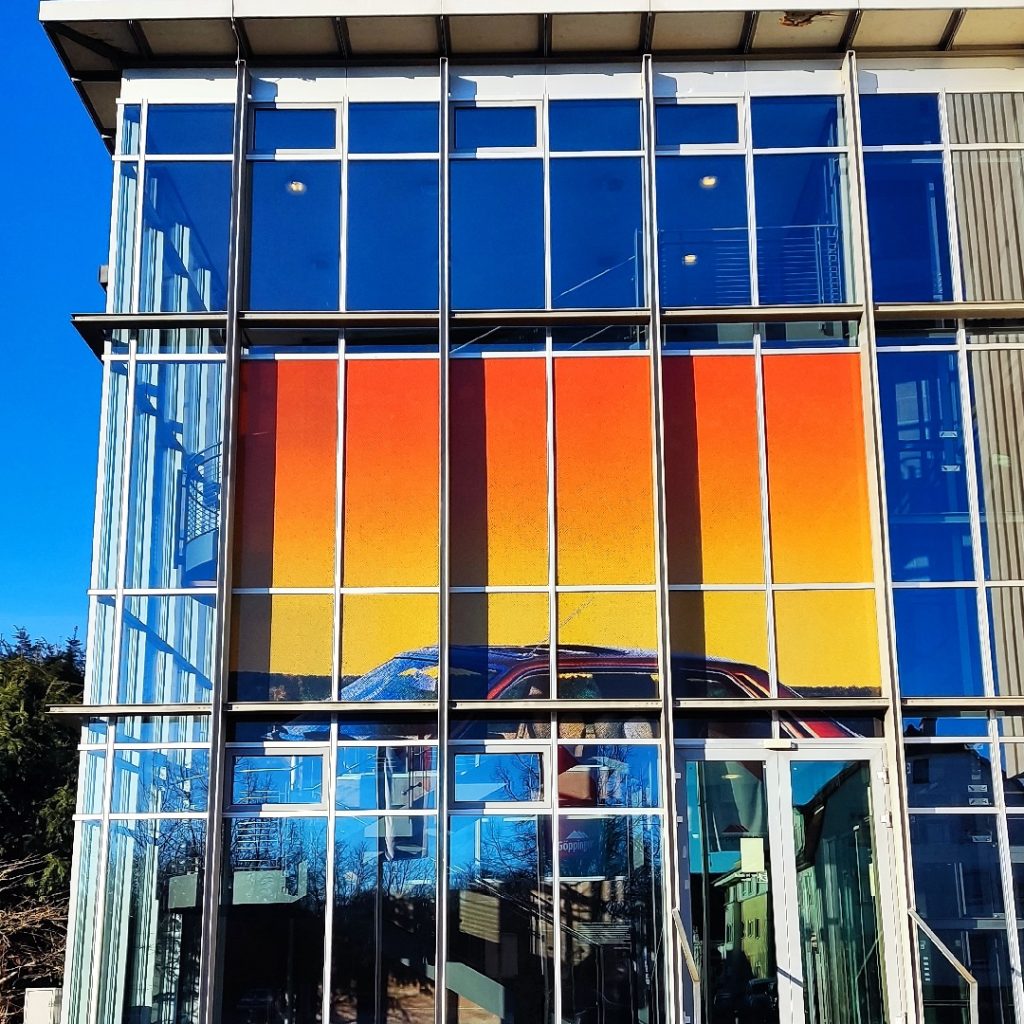 Glasfassade mit dem Kunstwerk GOLDEN von Simone Demandt: Farbige Streifen in Rot, Orange und Gelb, klare Linien und Spiegelungen bei sonnigem Wetter.