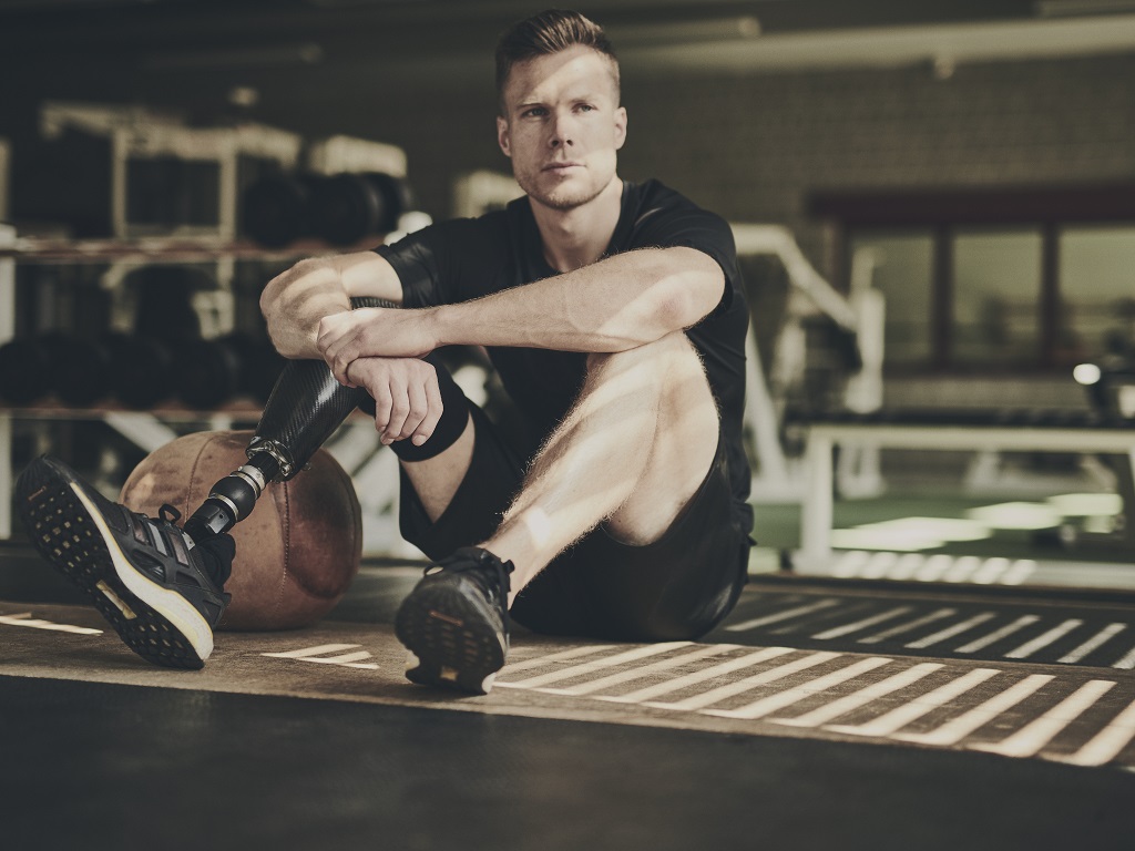 Markus Rehm sitzt in Trainingskleidung auf dem Boden eines Fitnessstudios, den Rücken leicht angelehnt an einen Medizinball. Sein linker Unterschenkel ist amputiert und durch eine Prothese ersetzt. Er blickt ruhig und nachdenklich in die Ferne.