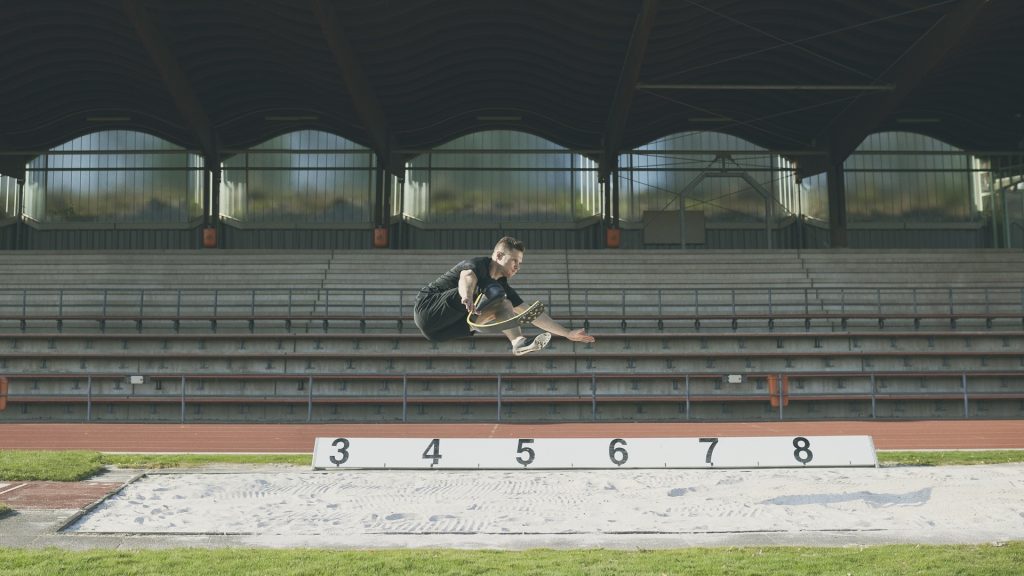 Markus Rehm, ein Para-Weitspringer mit Prothese am linken Bein, springt weit über eine Weitsprunggrube in einem leeren Stadion. Er trägt Sportkleidung und befindet sich in der Luft in kompakter Haltung, vor leeren Tribünen im Hintergrund.