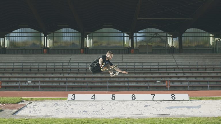 Markus Rehm, ein Para-Weitspringer mit Prothese am linken Bein, springt weit über eine Weitsprunggrube in einem leeren Stadion. Er trägt Sportkleidung und befindet sich in der Luft in kompakter Haltung, vor leeren Tribünen im Hintergrund.