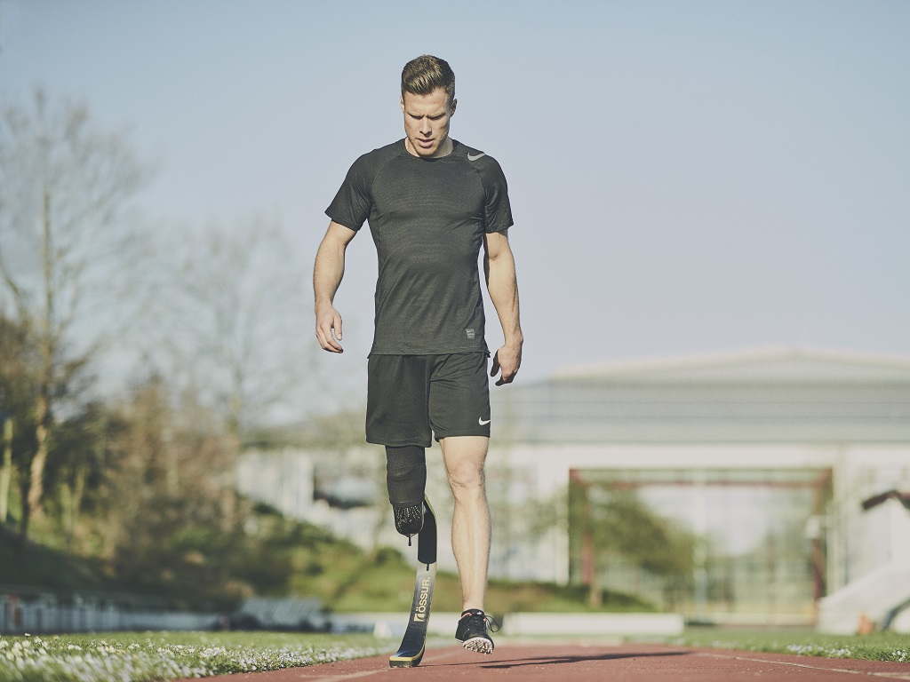 Markus Rehm, Para-Athlet mit Unterschenkelprothese, geht im Trainingsoutfit über eine Laufbahn. Er blickt konzentriert nach unten, im Hintergrund sind Bäume und ein modernes Gebäude zu sehen.