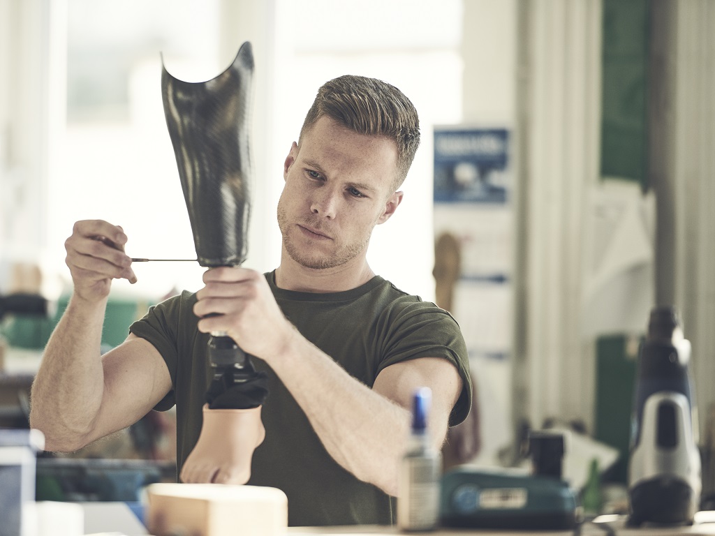Markus Rehm arbeitet konzentriert an einer Beinprothese in einer Werkstatt. Er hält das Modell mit beiden Händen und justiert es mit einem Werkzeug – umgeben von typischen Arbeitsmaterialien.