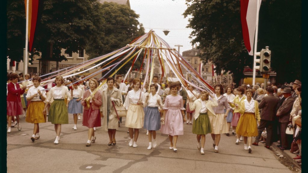 Festlich gekleidete Mädchen führen beim Göppinger Maientag 1961 eine traditionelle Bändertanz-Gruppe an. Sie halten farbige Bänder, die an einer zentralen Spitze befestigt sind, während sie durch die geschmückte Innenstadt ziehen – umgeben von zahlreichen Zuschauern. Das historische Farbfoto vermittelt lebendige Eindrücke eines beliebten Brauches in der Stadtgeschichte.