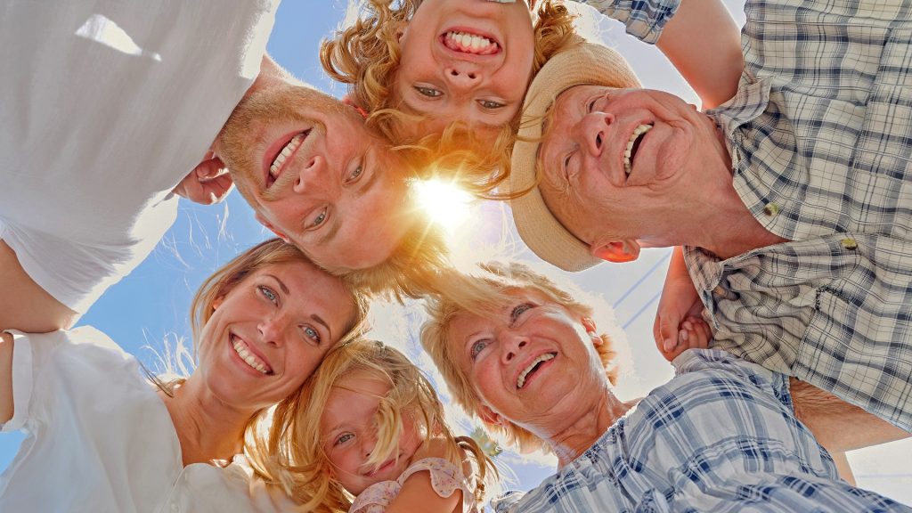 Sechs fröhliche Menschen aus drei Generationen – Kinder, Eltern und Großeltern – stehen im Kreis und blicken lachend in die Kamera, mit Sonnenschein im Hintergrund. Symbolbild für generationsübergreifende Verbundenheit und Vorsorge.