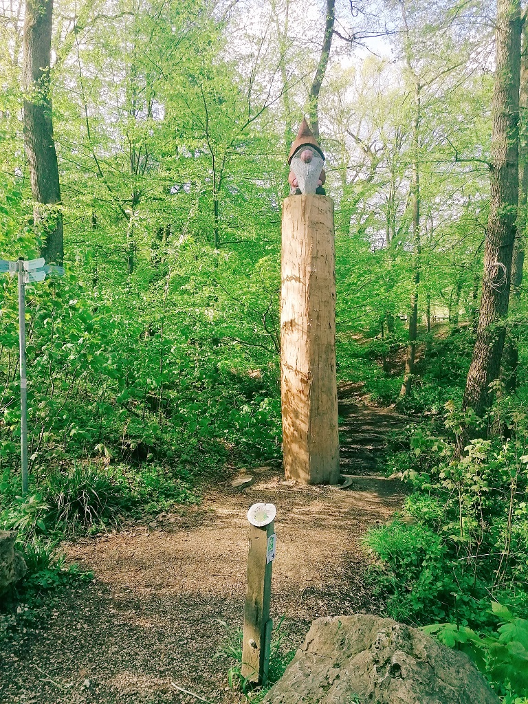 Waldweg mit hoher Holzsäule und Zwergenfigur.