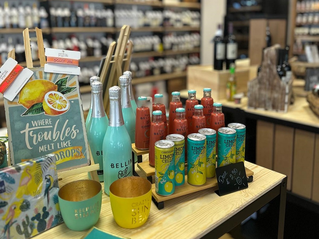 Nahaufnahme eines Präsentationstisches bei Solo Vino mit bunten Limoncello-Dosen, Aperitifflaschen, Sommeraccessoires und Dekoartikeln, im Hintergrund Weinregale.