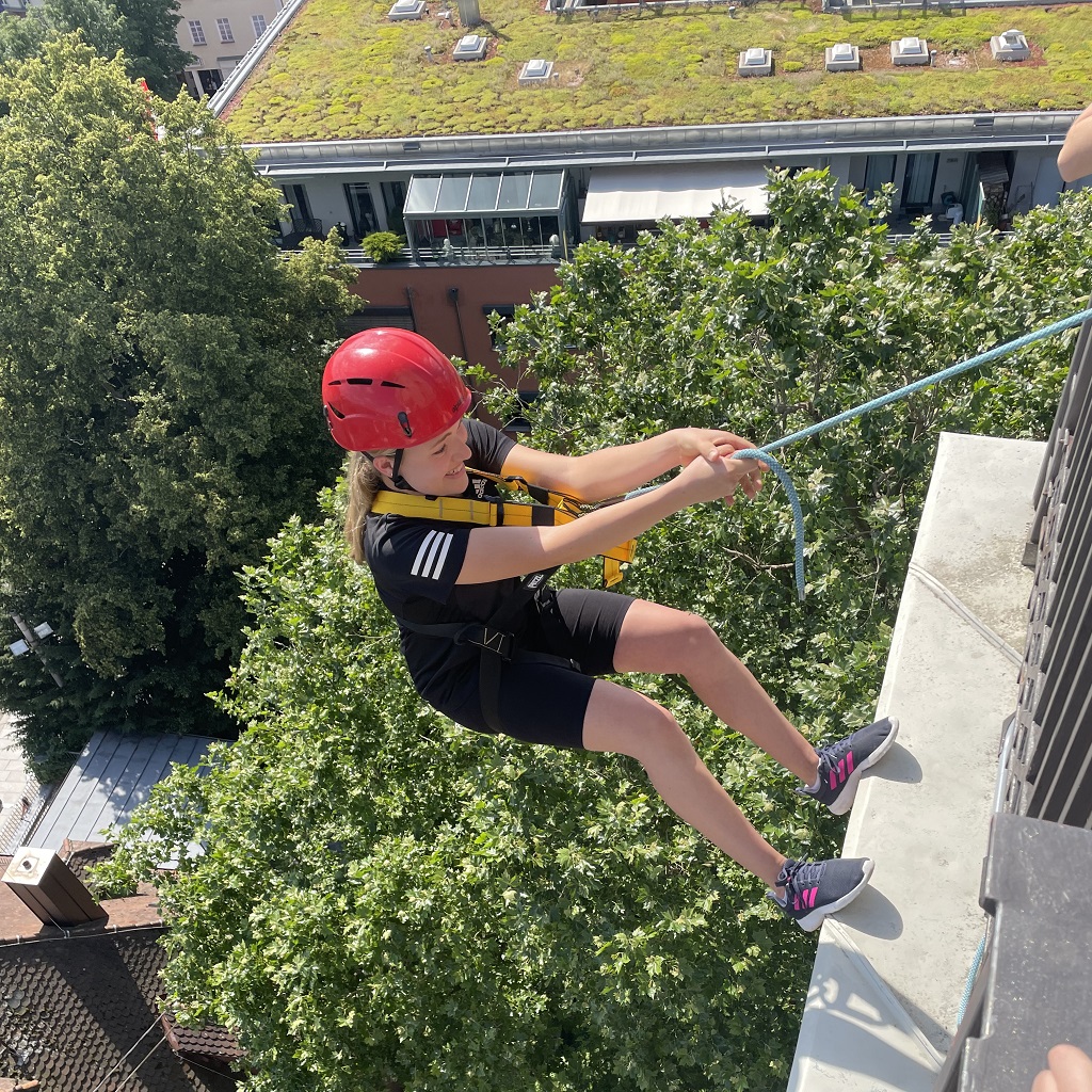 Jugendliche mit Klettergurt und Helm beim Abseilen vom Turm der Stadtkirche Göppingen – mit Blick auf Baumwipfel und Dächer der Stadt.