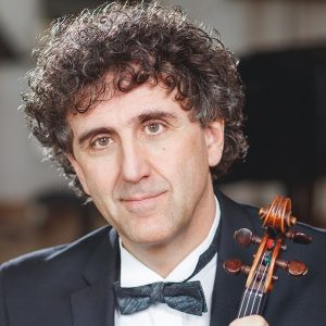 Porträt von Stadtmusikdirektor Martin Gunkel im Anzug mit Fliege, eine Violine in der Hand haltend, Blick in die Kamera.