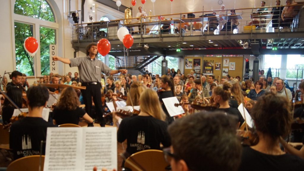 Dirigent und Jugendsinfonieorchester der Jugendmusikschule Göppingen beim Kinderkonzert im Alten E-Werk, mit vielen zuschauenden Kindern und Familien im Publikum, geschmückt mit roten Luftballons.