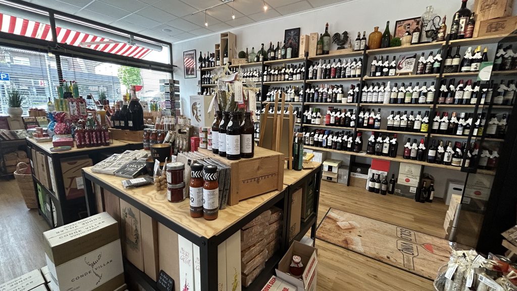 Innenaufnahme des Solo Vino Weinhandels in Ebersbach: Große Auswahl an Weinflaschen in Wandregalen, im Vordergrund Tische mit Feinkost, Saucen, Geschenkartikeln und dekorativen Produkten, durch große Fenster fällt Tageslicht.
