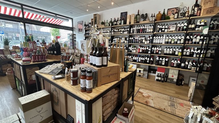 Innenaufnahme des Solo Vino Weinhandels in Ebersbach: Große Auswahl an Weinflaschen in Wandregalen, im Vordergrund Tische mit Feinkost, Saucen, Geschenkartikeln und dekorativen Produkten, durch große Fenster fällt Tageslicht.