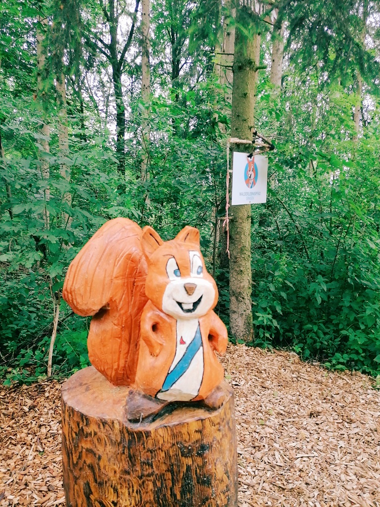 Holzfigur eines lachenden Eichhörnchens auf einem Baumstamm am Walderlebnispfad Gingen, mit Wegweiser im Hintergrund inmitten eines grünen Waldes.